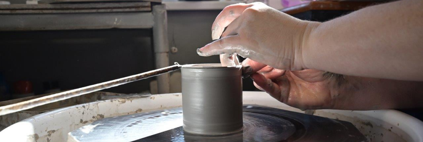 Detail rukou pracujících na hrnčířském kruhu, uprostřed kterého je základ hrnku.